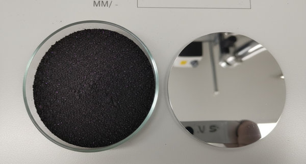 Dark NMC cathode powder in a glass dish beside a reflective circular cobalt sputtering target disc