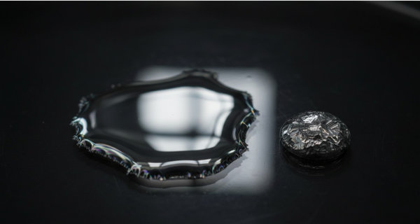 Reflective pool of liquid gallium metal with iridescent edges beside a crystalline solidified droplet on dark glass
